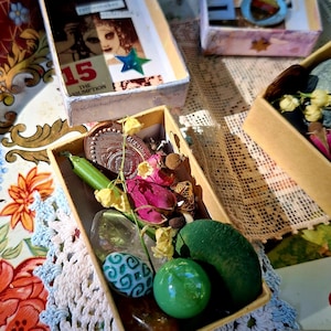 May include: A rectangular cardboard box filled with a collection of small items including a copper coin, a green marble, a dried pink rose, a green plastic pea pod, a smooth green stone, a clear crystal, and a small white bead with blue patterns.