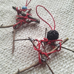 May include: Two handmade twig ornaments. Each ornament is crafted from small branches, red twine, and silver wire. One features a blue bead, the other a dark, textured sphere. Both have red hanging strings.