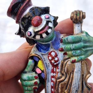 May include: A hand-painted clown figurine with a green face, black hair, and a red nose. The clown wears a top hat and holds a violin. The figurine has a colorful outfit with red, white, blue, yellow, and pink accents.