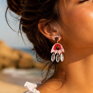 Puede incluir: Un pendiente colgante con un poste en forma de ojo rosa con un centro oscuro, conectado a un dije más grande en forma de ojo rosa con iris rojo y pupila oscura. Tres cristales transparentes, facetados y en forma de marquesa cuelgan.