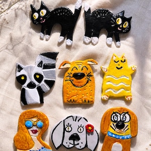 Puede incluir: Un conjunto de ocho broches de cerámica pintados a mano con varios personajes, incluyendo dos gatos negros con patas blancas, un lémur, una criatura amarilla, una persona con gafas de sol, un perro blanco con una flor roja y un perro marrón.