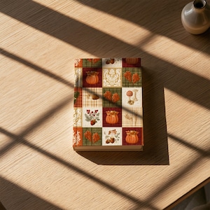 May include: A rectangular book with a patchwork cover design featuring autumn-themed illustrations. The grid pattern displays orange pumpkins, red and brown leaves, acorns, berries, mushrooms, and white floral motifs against green, red, and plaid backgrounds.