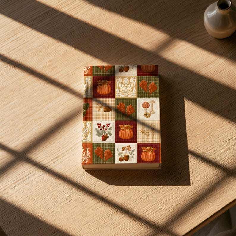 May include: A rectangular book with a patchwork cover design featuring autumn-themed illustrations. The grid pattern displays orange pumpkins, red and brown leaves, acorns, berries, mushrooms, and white floral motifs against green, red, and plaid backgrounds.