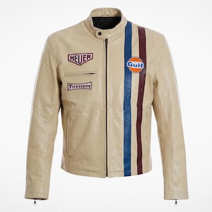 May include: A beige leather jacket featuring a central zipper, a mandarin collar with a snap button, and two vertical stripes in blue and maroon. The chest displays embroidered patches with the text Heuer, Firestone, and a circular Gulf logo.