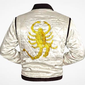 May include: A quilted off-white satin bomber jacket featuring a large, embroidered yellow scorpion on the back. The jacket has a dark brown ribbed collar, cuffs, and hem, with long sleeves and a diamond-stitched pattern across the entire back panel.