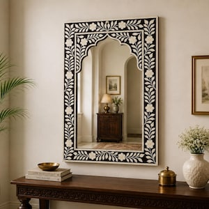 May include: A rectangular wall mirror featuring a dark brown frame with an intricate white floral and vine inlay pattern. The mirror has a decorative arched top design and is mounted on a plain wall above a wooden console table.