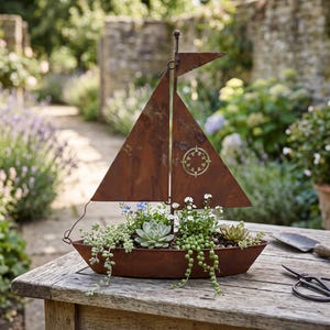 Puede incluir: Una maceta de metal color óxido con forma de velero que presenta una vela triangular con un diseño de rosa de los vientos recortado. El casco del barco está lleno de varias suculentas verdes y pequeñas flores blancas decorativas.