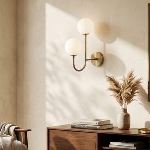 Puede incluir: Un aplique de pared con estructura de metal en tono latón y un brazo curvo que sostiene dos globes esféricos de vidrio blanco. La lámpara se fija a la pared mediante una placa base circular en tono latón, proporcionando iluminación.