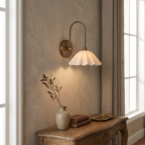May include: A wall-mounted light fitting featuring a curved brass arm and a white, scalloped, bell-shaped glass shade. The lamp is attached to a circular brass wall plate and emits a soft, downward glow, illuminating the wall surface below it.