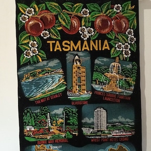 May include: A black fabric wall hanging featuring the text TASMANIA, illustrations of fruit, and five scenes: The Nut at Stanley, Ulverstone, Princes Square Fountain Launceston, Burnie War Memorial, and Wrest Point Hotel Casino, with a map at the bottom.