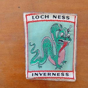 May include: A rectangular patch with a green cartoon Loch Ness Monster, pink belly, and red tongue. The text "LOCH NESS" and "INVERNESS" are in white on a red and white border. The patch is on a light green background.
