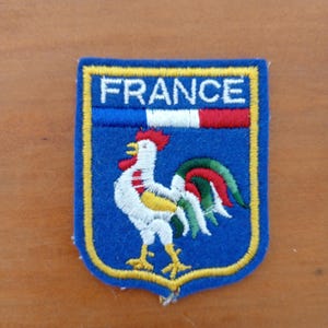 May include: A blue embroidered patch with a yellow border and the word "FRANCE" at the top. The patch features a white rooster with red, yellow, and green details, a symbol of France.
