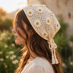 May include: A cream-colored crochet headscarf featuring a repeating pattern of white daisy appliques with yellow centers. The headscarf is constructed with an open-knit texture and includes two braided ties at the back, each finished with a tassel.