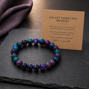 Könnte beinhalten: Ein Perlenarmband mit polierten, kugelförmigen Galaxy Tiger's Eye-Steinen in tiefen Violett-, Blau- und Petroltönen. Daneben liegt eine beigefarbene Karte mit dem Text: GALAXY TIGER'S EYE BRACELET. Wear this bracelet as a reminder of your inner strength and limitless potential. May it bring you courage, confidence, and clarity on your journey. You are capable of amazing things. Believe in yourself.