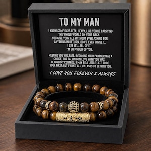 Könnte beinhalten: Eine schwarze Geschenkbox mit einem Perlenarmband aus braunen Tigeraugen-Steinen, einer goldfarbenen zylindrischen Perle mit schwarzen Akzenten und einer goldfarbenen Kugelperle mit schwarzen Steinen. Der Deckel trägt den Text TO MY MAN und einen Text.