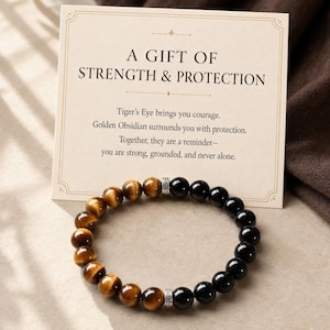 May include: A beaded bracelet featuring alternating sections of brown tiger's eye and black obsidian stones, accented by two silver-toned metal spacers with a Greek key pattern. The bracelet is displayed in front of a card with the text A GIFT OF STRENGTH & PROTECTION.