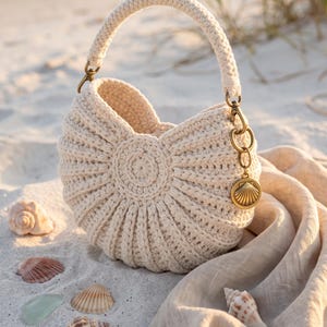 Pode incluir: Uma bolsa de crochê na cor creme com um design arredondado e canelado, com uma alça única. A bolsa apresenta um fecho de metal em tom de latão na lateral, que sustenta um pingente circular gravado com um motivo de concha marinha.