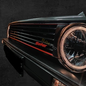 Könnte beinhalten: Ein schwarzer Autokühlergrill mit einer horizontalen roten Akzentlinie und dem Text Gofer SLOA.PRO. Die Baugruppe umfasst einen großen runden Scheinwerfer mit reflektierendem Innengehäuse und ein rechteckiges bernsteinfarbenes Blinklicht unter der Stoßstange.