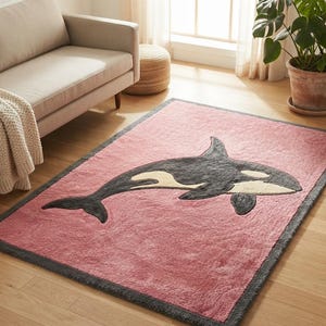 Puede incluir: Una alfombra rectangular con un campo rosa y un borde gris oscuro. Centrado en la alfombra hay un gráfico grande y estilizado de una orca en tonos negro, gris oscuro y crema, representado en movimiento de nado sobre la superficie rosa.
