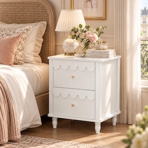 May include: A white wooden nightstand featuring two drawers with scalloped edges and small gold knobs. The piece stands on four turned legs and has a smooth, rectangular top surface. The design is clean and functional, providing storage space.