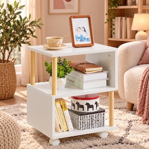 May include: A white two-tier side table with light wood cylindrical supports and four caster wheels. The top surface holds a beige ceramic cup on a saucer and a wooden picture frame. The shelves contain a potted plant, books, and storage boxes.