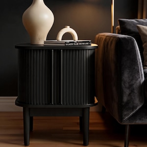 May include: A black bedside table with a tambour door front and four legs. On top sits a beige vase, a small beige arch-shaped sculpture, and a book titled Vanity Fair 100 Years. The table features vertical slats and two thin vertical handles.