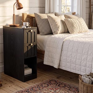 May include: A tall, narrow black nightstand with two drawers featuring vertical grooved detailing and gold-toned circular knobs. The unit includes an open bottom shelf, a flat top surface holding a copper-colored desk lamp, and a white ceramic mug.