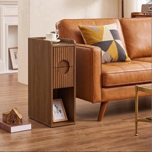 May include: A tall, narrow wooden side table with a dark brown finish, featuring a top surface, a single drawer with a vertical fluted texture and a circular cutout handle, and an open bottom shelf containing a small framed picture of a cactus.