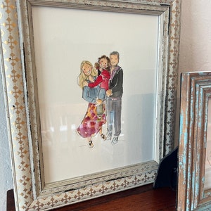 May include: A framed watercolor portrait of a family, featuring a woman, a child, and a man. The artwork is in a distressed white and gold frame. The family is depicted in casual attire, with the woman wearing a floral skirt.