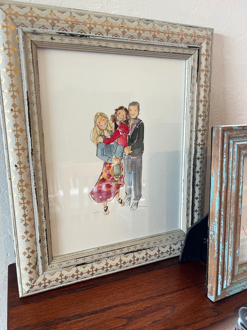 May include: A framed watercolor portrait of a family, featuring a woman, a child, and a man. The artwork is in a distressed white and gold frame. The family is depicted in casual attire, with the woman wearing a floral skirt.