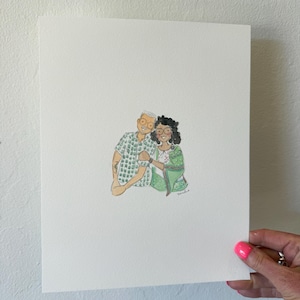 May include: Watercolor portrait of a couple, featuring a man in a green patterned shirt and a woman in a green sari. The artwork is on white paper, held by a hand with pink nail polish.