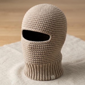 Op de afbeelding: Een beige gehaakte bivakmuts met een grote opening voor de ogen en een geribbelde halskraag. Het hoofddeksel is gemaakt met een dicht, uniform steekpatroon en bevat een klein rechthoekig stoffen label aan de onderkant met de letters LK.