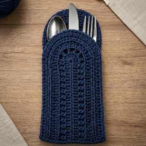 Puede incluir: Un porta cubiertos de ganchillo en color azul oscuro con un borde superior redondeado, que contiene una cuchara, un cuchillo y un tenedor de acero inoxidable. El soporte presenta una textura acanalada vertical y un patrón semicircular.