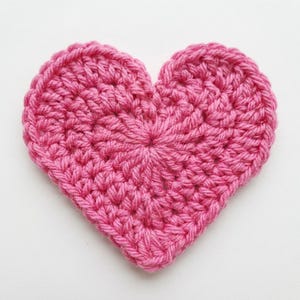 May include: A heart-shaped crochet applique crafted from vibrant pink yarn. The piece features a dense, textured stitch pattern radiating from the center, forming a clear heart silhouette with a rounded top and a pointed bottom edge.