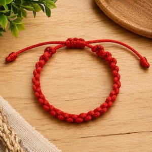 May include: A bright red cord bracelet featuring a continuous series of intricate, hand-knotted loops. The design includes an adjustable sliding knot closure with two thin cord ends, each finished with a small, secure knot at the very tip.