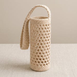 May include: A cylindrical beige crochet bottle holder featuring an open-work grid pattern and a single integrated handle. The structure is crafted from thick yarn, creating a sturdy, hollow vessel designed to carry a bottle with a visible open weave.