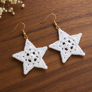 Può includere: Un paio di orecchini a forma di stella bianca, realizzati con un intricato filo all'uncinetto. Ogni stella presenta un motivo centrale traforato ed è appesa a un gancio in metallo dorato. Gli orecchini sono appoggiati su legno scuro.