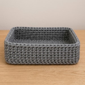 May include: A rectangular storage basket crafted from thick, dark gray yarn. The basket features a consistent, dense crochet stitch pattern throughout its walls and base, creating a structured, open-top container with a uniform, textured appearance.