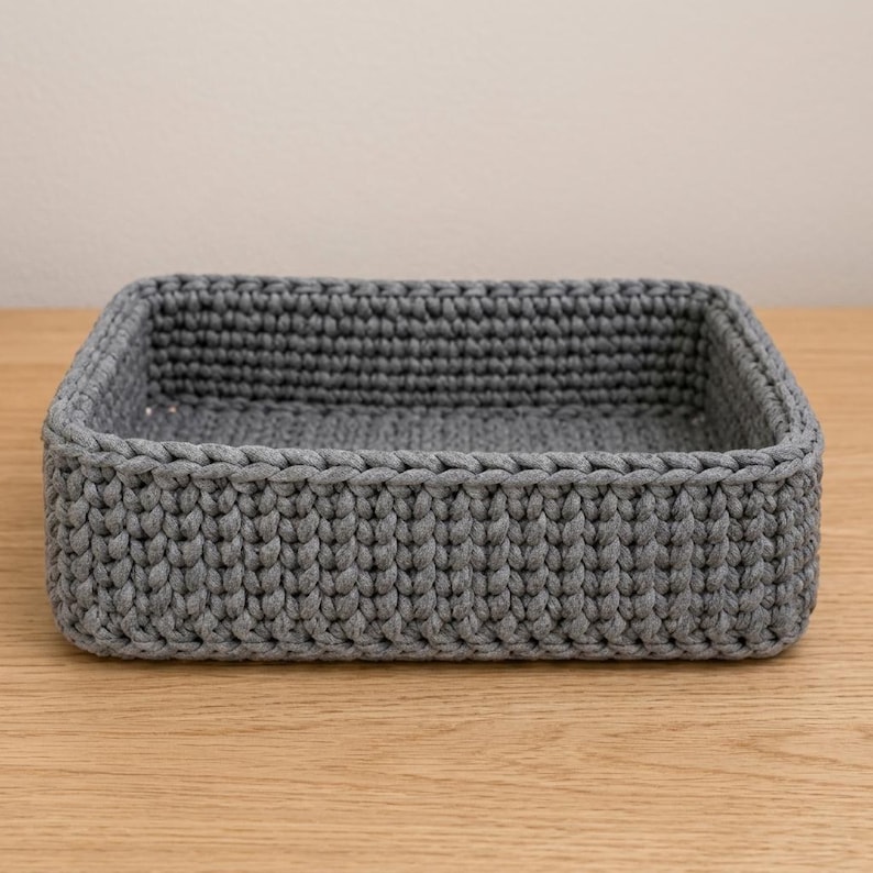 May include: A rectangular storage basket crafted from thick, dark gray yarn. The basket features a consistent, dense crochet stitch pattern throughout its walls and base, creating a structured, open-top container with a uniform, textured appearance.