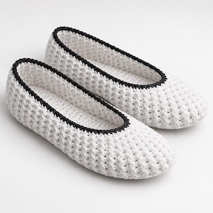 Puede incluir: Un par de zapatillas de ganchillo blancas con un patrón de puntos texturizados tipo gofre en todo el cuerpo. Cada zapatilla está rematada con un borde de ganchillo negro sólido a lo largo de la abertura, creando un contraste con el hilo blanco.