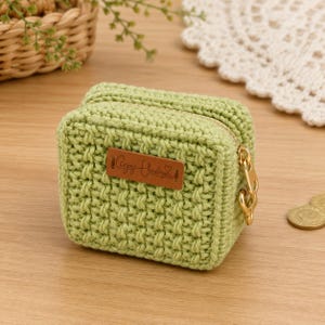 May include: A small, rectangular, light green crochet pouch with a textured stitch pattern. It features a gold-tone metal zipper closure and a rectangular brown leather-like label on the front with the text Enjoy Handmade printed in black.