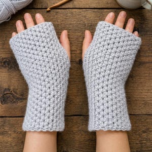 May include: A pair of light gray fingerless gloves featuring a textured crochet stitch pattern. The gloves cover the hand from the knuckles down to the wrist, leaving the fingers and thumb exposed, showcasing a consistent knit-like construction.