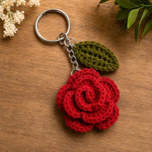 May include: A keychain featuring a crocheted red rose flower and a single crocheted olive green leaf attached to a silver metal split ring by a short silver chain. The rose has layered, textured petals and the leaf has a visible stitched pattern.