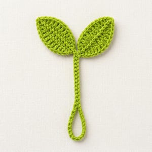 May include: A bright lime green crochet applique shaped like two leaves attached to a central stem that forms a small loop at the bottom. The leaves feature a visible textured stitch pattern with a central vein detail, crafted from yarn.