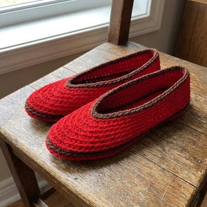 Peut inclure: Une paire de chaussons en crochet rouge vif avec une bordure marron foncé le long du col et une bande horizontale marron assortie entourant la base de la chaussure. Les chaussons présentent un bout arrondi et une ouverture basse.