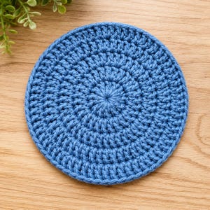 May include: A circular blue coaster crafted with a dense crochet stitch pattern. The textile item features a consistent, repeating yarn texture that radiates from the center outward to the finished edge, creating a uniform and flat circular shape.