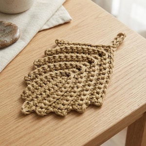 May include: A tan, leaf-shaped crochet coaster made from thick yarn. The coaster features a textured, ribbed stitch pattern that mimics the veins of a leaf, with a small braided loop at the top point for hanging. The item is a solid, neutral colour.