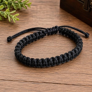 Peut inclure: Un bracelet en cordon noir présentant un motif en macramé tressé avec une fermeture à nœud coulissant. Le bracelet est fabriqué à partir d'un cordon noir continu, avec deux extrémités réglables se terminant par de petits nœuds aux extrémités.