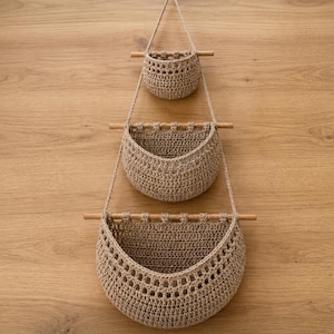 May include: A three-tiered hanging wall organizer featuring three beige crocheted baskets of varying sizes. Each basket is suspended by thin cords from a horizontal wooden dowel, creating a vertical arrangement for storage and organization.