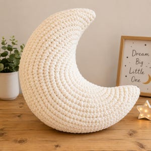 May include: A crescent moon-shaped pillow crafted from off-white yarn with a visible crochet stitch pattern. The pillow has a curved, three-dimensional form and a soft, textured surface, standing upright on a wooden surface.